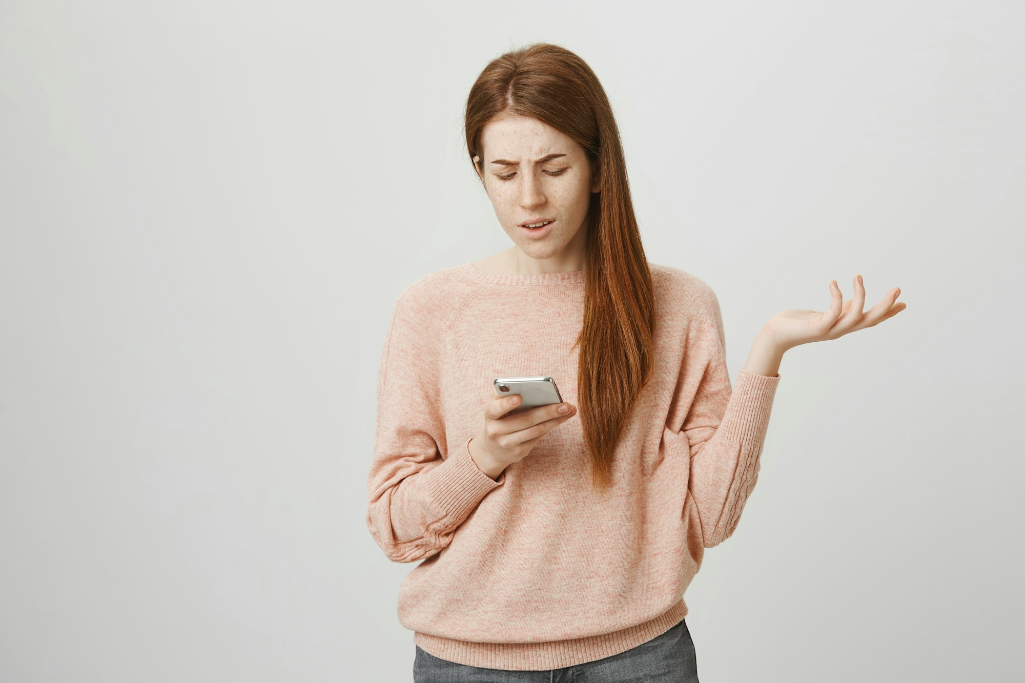 Une jeune fille rousse inquiète ou irritée gesticule en regardant l'écran d'un smartphone.