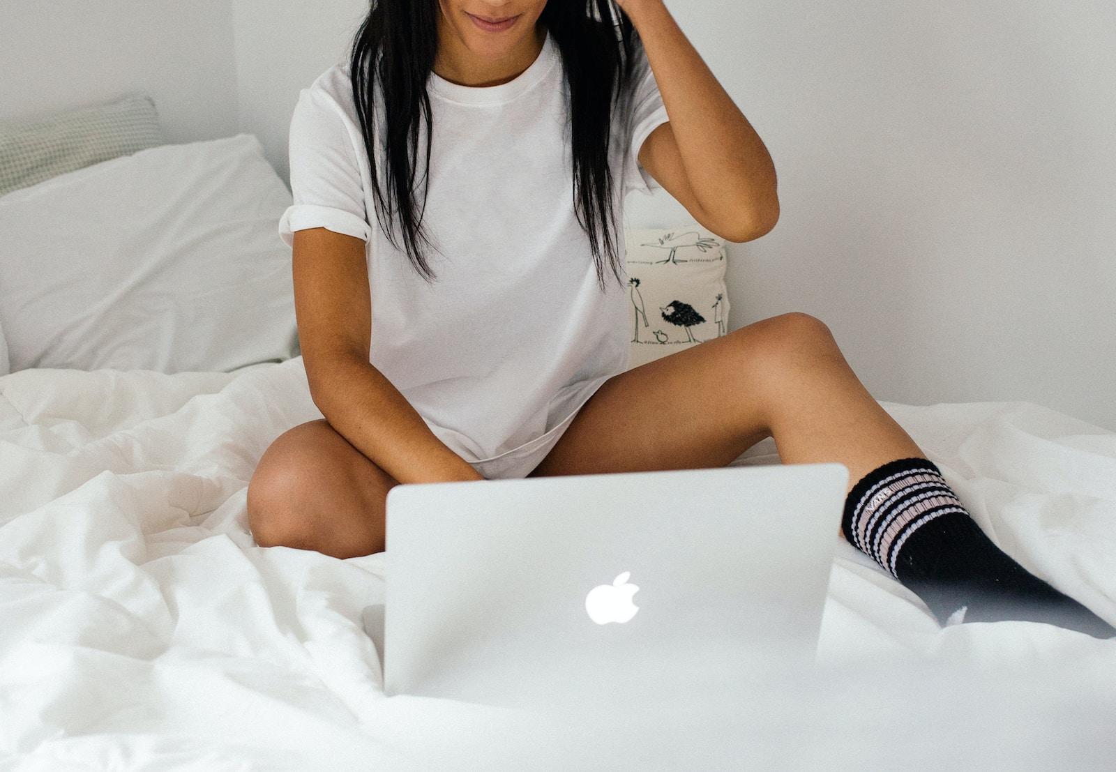 Une femme en t-shirt blanc sur un lit, tenant un MacBook argenté.