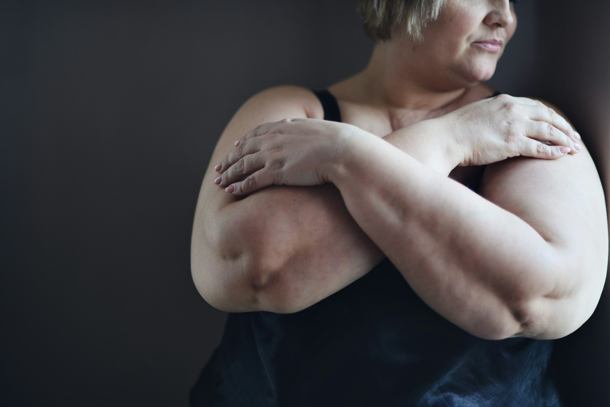 Une grosse femme déprimée se tient debout, les bras croisés, et regarde par la fenêtre de sa maison.