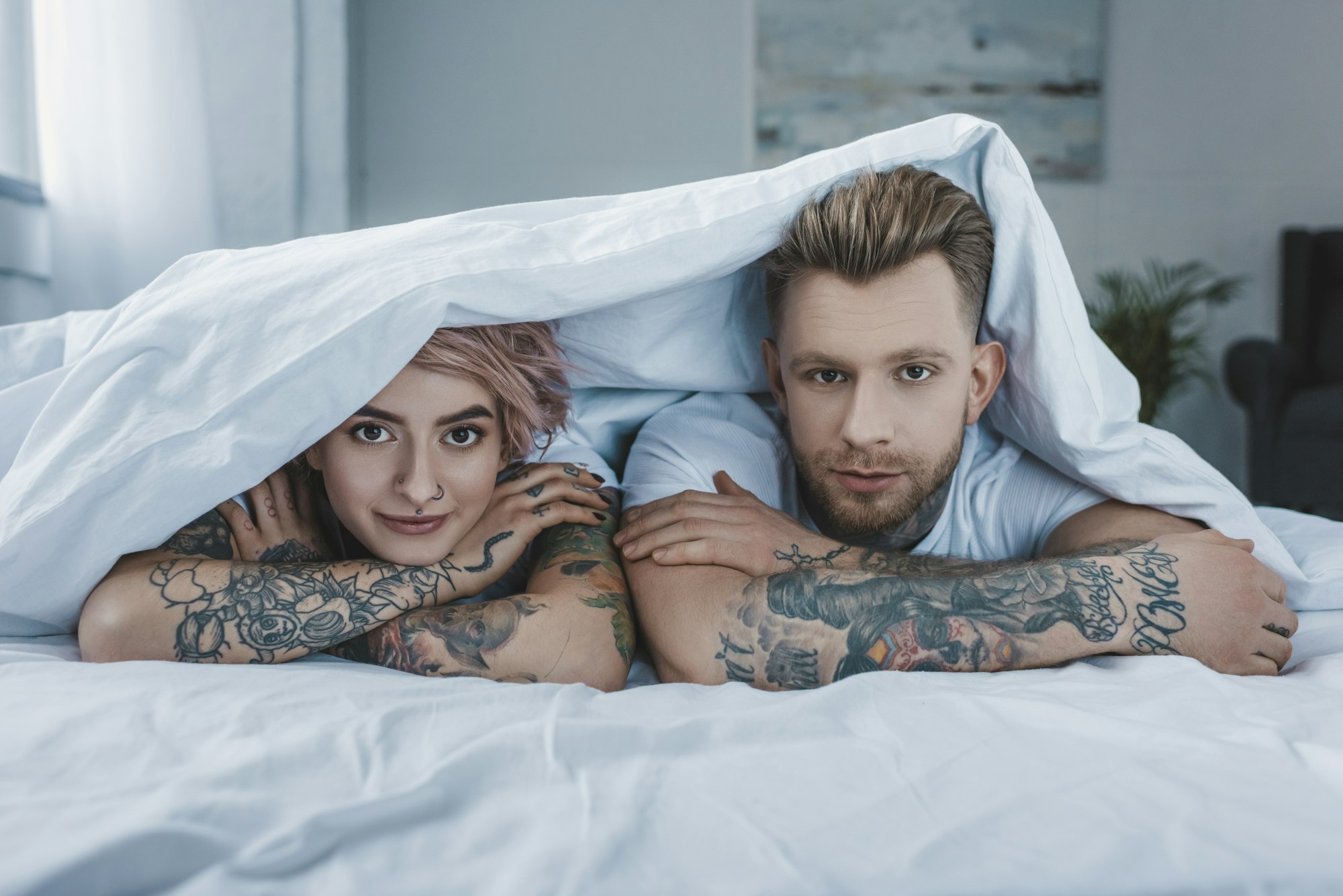 jeune couple tatoué allongé dans un lit sous une couverture blanche