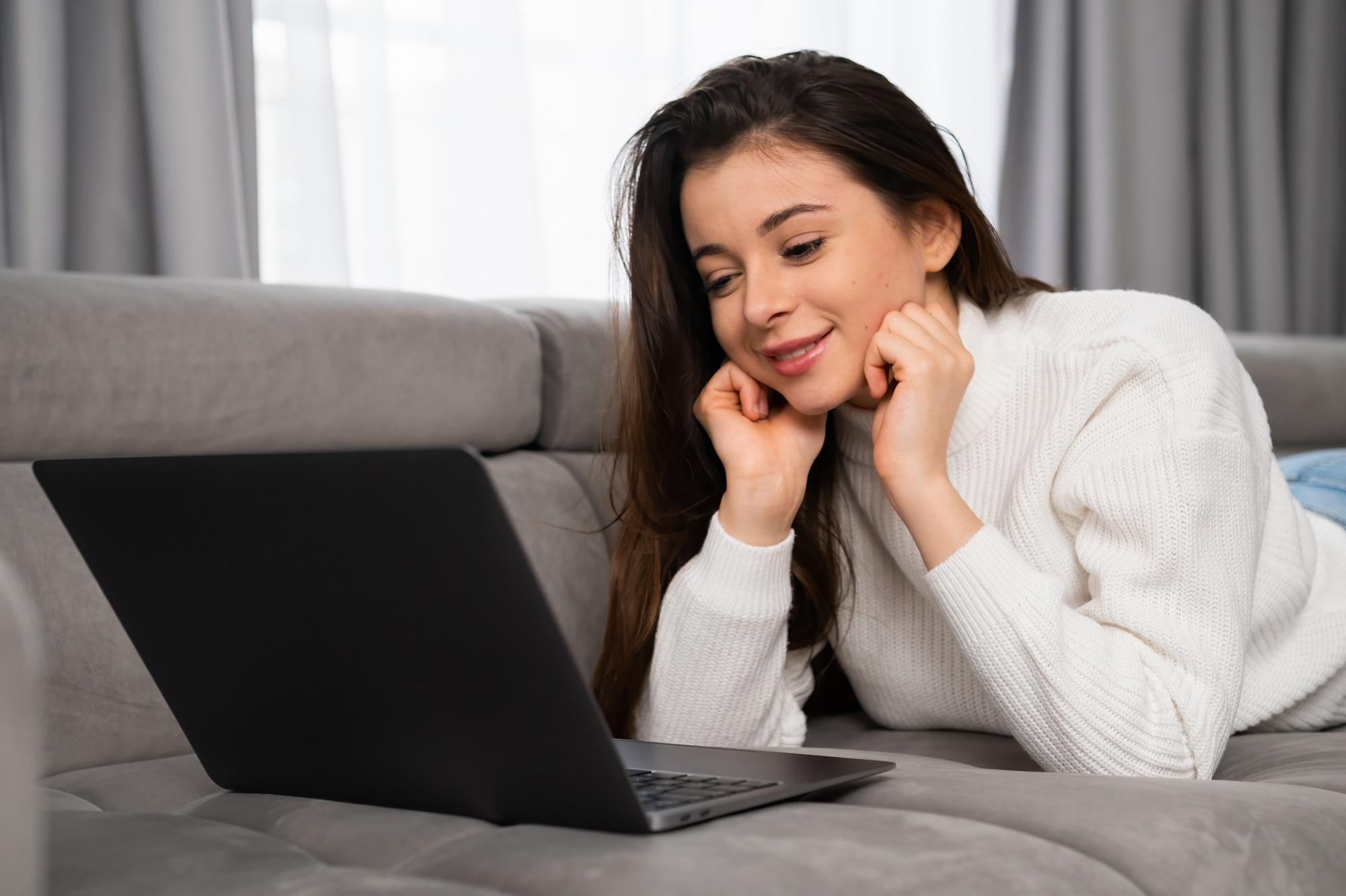 Jeune fille souriante lors d'un rendez-vous en ligne via un ordinateur portable, allongée sur un canapé dans son appartement.