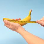 Hands measuring banana with measuring yellow tape on blue background